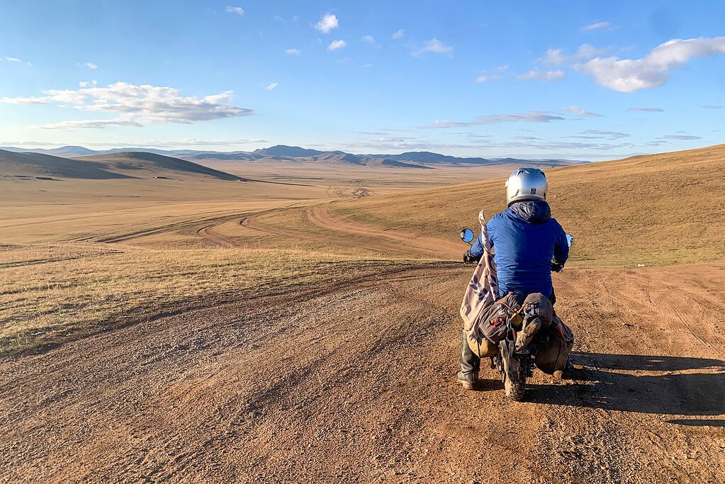 A loaded monkey bike catching air off a grass ridge on the Mongolian steppe